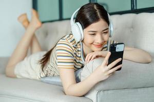 bella donna asiatica sdraiata sul divano e ascoltando musica mentre usa il telefono