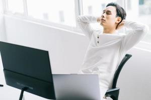 il programmatore asiatico si siede con la testa all'indietro sulla sedia per rilassarsi dopo un faticoso orario di lavoro foto