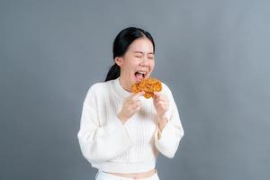 giovane donna asiatica che indossa un maglione con una faccia felice e si diverte a mangiare pollo fritto su sfondo grigio foto