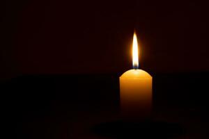 singolo ardente candela fiamma o leggero raggiante su un' grande giallo candela su nero o buio sfondo su tavolo nel Chiesa per Natale, funerale o memoriale servizio con copia spazio. foto