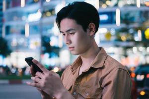 il giovane asiatico sta usando il suo telefono mentre cammina per strada di notte