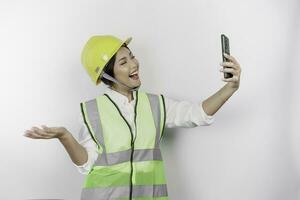 un' sorridente asiatico donna lavoro duro e faticoso indossare sicurezza casco e veste, Tenere sua Telefono e puntamento per copia spazio accanto suo, isolato di bianca sfondo. lavoro giorno concetto. foto