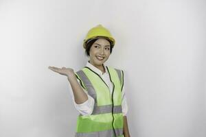 un' sorridente asiatico donna lavoro duro e faticoso indossare sicurezza casco e veste, puntamento per copia spazio accanto suo, isolato di bianca sfondo. lavoro giorno concetto. foto