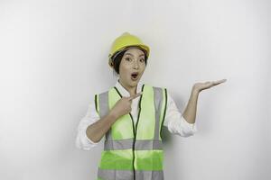 un' scioccato asiatico donna lavoro duro e faticoso indossare sicurezza casco e veste, puntamento per copia spazio accanto suo, isolato di bianca sfondo. lavoro giorno concetto. foto