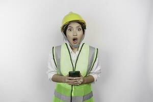 un' ritratto di un' scioccato asiatico donna lavoro duro e faticoso indossare sicurezza casco e veste mentre Tenere sua Telefono con sua bocca largo aprire, isolato di bianca sfondo. lavoro giorno concetto foto