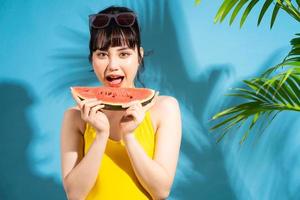 bella donna asiatica che indossa una tuta gialla su sfondo blu e mangia frutta tropicale, concetto estivo foto