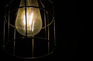 notte fotografia di il leggero lampadina, incandescente lampada, un' filo filamento quello è riscaldato fino a esso si illumina. con metallo gabbia isolato su nero sfondo. foto