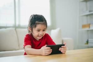 asiatico bambino ragazza guardare utilizzando e toccare mobile Telefono schermo. bambino sorridente divertente tempo per uso mobile Telefono. pure tanto schermo volta. carino ragazza Guardando video mentre tv nel vivente camera, Internet dipendenza. foto