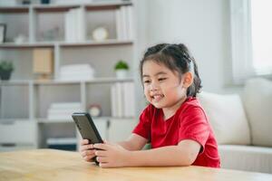 asiatico bambino ragazza guardare utilizzando e toccare mobile Telefono schermo. bambino sorridente divertente tempo per uso mobile Telefono. pure tanto schermo volta. carino ragazza Guardando video mentre tv nel vivente camera, Internet dipendenza. foto