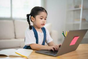 asiatico bambino ragazza sorridente uso il computer portatile o scrittura disegno colore su Nota libro studia in linea su legna tavolo scrivania nel vivente camera a casa. formazione scolastica apprendimento in linea a partire dal casa concetto. foto