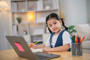 asiatico bambino ragazza sorridente uso il computer portatile o scrittura disegno colore su Nota libro studia in linea su legna tavolo scrivania nel vivente camera a casa. formazione scolastica apprendimento in linea a partire dal casa concetto. foto