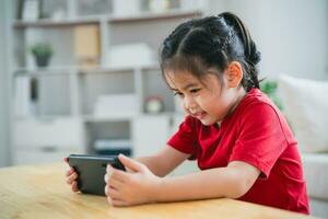 asiatico bambino ragazza guardare utilizzando e toccare mobile Telefono schermo. bambino sorridente divertente tempo per uso mobile Telefono. pure tanto schermo volta. carino ragazza Guardando video mentre tv nel vivente camera, Internet dipendenza. foto