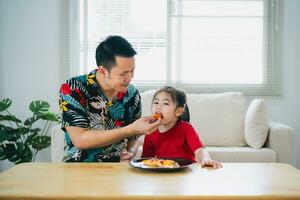 asiatico contento bambini ragazza e sua padre alimentazione mangiare Pizza e sorridente nel il vivente camera a casa. bambini ragazza e sua papà mangiare e degustazione italiano fatti in casa Pizza nel Casa. foto