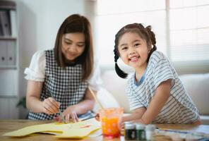 carino poco bambino pittura con colorato vernici. asiatico ragazza e madre utilizzando pastello disegno colore. figlia e mamma fare compiti a casa colorazione cartone animato personaggi. bambino artista attività stile di vita concetto. foto