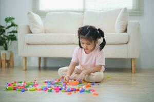 gioioso asiatico ragazza contento e sorridente giocando colorato Lego giocattoli, seduta su il vivente camera pavimento, creativamente giocando con Lego, edificio colorato strutture creatività immaginare. apprendimento formazione scolastica. foto