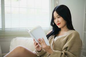 gioioso contento asiatico donna sorridente e lettura libro mentre seduta su divano divano nel vivente camera a casa. asiatico donne rilassare lettura libro Sorridi a divano nel il Casa. attività passatempo a Casa concetto. foto