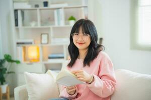 gioioso contento asiatico donna sorridente e lettura libro mentre seduta su divano divano nel vivente camera a casa. asiatico donne rilassare lettura libro Sorridi a divano nel il Casa. attività passatempo a Casa concetto. foto