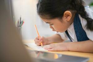 asiatico bambino ragazza sorridente uso il computer portatile o scrittura disegno colore su Nota libro studia in linea su legna tavolo scrivania nel vivente camera a casa. formazione scolastica apprendimento in linea a partire dal casa concetto. foto