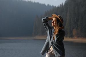 la giovane donna felice in cappello gode della vista del lago nella foresta. momenti di relax. vista della ragazza alla moda gode della freschezza all'aperto. libertà, persone, stile di vita, viaggi e vacanze foto