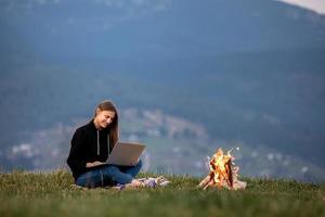 giovane donna freelance con laptop in montagna la sera. ragazza turistica seduta vicino al fuoco