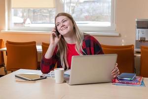 una donna bionda sorridente seduta a un computer portatile e parlando al cellulare. foto