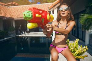 bella sottile donna giocando con pistola ad acqua giocattolo a piscina su estate tropicale vacanza foto