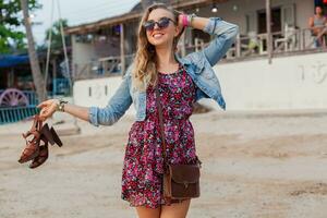 elegante donna nel estate vestito vacanza a piedi su spiaggia con scarpe nel mano foto