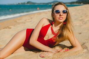 giovane bellissimo biondo donna prendere il sole su sabbia spiaggia nel rosso nuoto completo da uomo, occhiali da sole foto