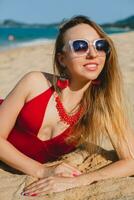 giovane bellissimo biondo donna prendere il sole su sabbia spiaggia nel rosso nuoto completo da uomo, occhiali da sole foto