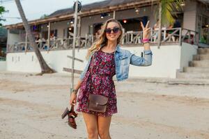 elegante donna nel estate vestito vacanza a piedi su spiaggia con scarpe nel mano foto