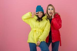Due attraente ragazza amici attivo donne in posa su rosa sfondo nel colorato inverno giù giacca di luminosa rosso e giallo colore avendo divertimento insieme, caldo cappotto moda tendenza, Cordiali saluti ridendo foto