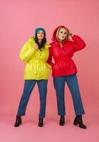 Due eccitato attraente ragazza amici attivo donne in posa su rosa sfondo nel colorato inverno giù giacca di luminosa rosso e giallo colore avendo divertimento insieme, caldo cappotto moda tendenza foto