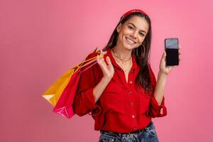 ispanico bellissimo donna nel rosso camicia sorridente Tenere Tenere shopping borse e smartphone foto