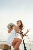 giovane attraente sorridente donna nel bianca vestito equitazione su tropicale spiaggia su bicicletta foto