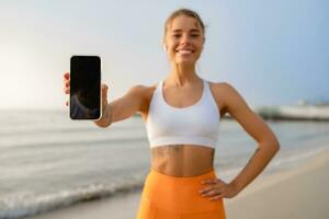 bella giovane sorridente donna fare gli sport nel il mattina nel elegante sport attrezzatura abbigliamento sportivo, Tenere smartphone schermo avvicinamento foto