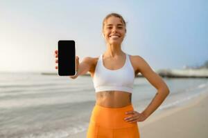 bella giovane sorridente donna fare gli sport nel il mattina nel elegante sport attrezzatura abbigliamento sportivo, Tenere smartphone schermo avvicinamento foto