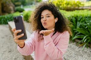 bella bellissimo Riccio giovane donna seduta nel parco utilizzando smatphone foto