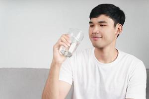 bell'uomo asiatico che beve un bicchiere d'acqua sul divano del soggiorno foto