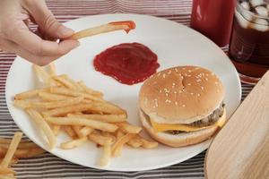 colpo del primo piano, messa a fuoco selettiva. la mano dell'uomo stava mangiando patatine fritte, immergendo il ketchup su un piatto bianco su una tovaglia rossa con hamburger e coca cola. mangiare cibo spazzatura o fast food per pranzo non è salutare. foto