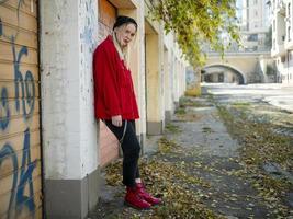 ragazza carina con un cappello a maglia, giacca rossa e stivali rossi in piedi vicino a un muro in piena crescita all'aperto foto