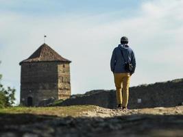 un uomo con un maglione con cappuccio e un cappello lavorato a maglia sta camminando lungo la strada per la fortezza sullo sfondo della torre e del cielo blu foto