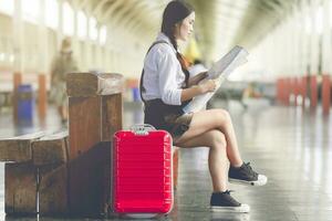 asiatico donna incinta sedersi su il panchina Guarda a il carta geografica con rosso valigia a ferrovia stazione viaggi, vacanze viaggio concetto. foto