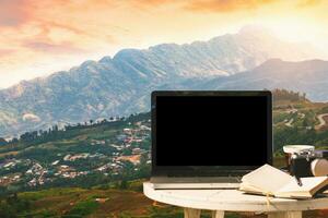 modello Immagine di il computer portatile con vuoto schermo con macchina fotografica, taccuino, caffè tazza su tavolo con a montani paesaggio contro tramonto cielo sfondo, tempo libero e viaggio concetto. foto