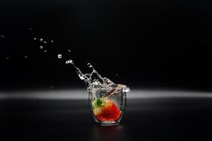 una fragola cade in un bicchiere di acqua limpida causando spruzzi d'acqua sullo sfondo nero foto