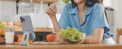 un' giovane donna con un' bellissimo viso nel un' blu camicia con lungo capelli mangiare frutta seduta dentro il cucina a casa con un' il computer portatile e taccuino per rilassamento, concetto vacanza. foto