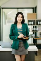 fiducioso asiatico donna con un' Sorridi in piedi Tenere bloc notes e tavoletta a il ufficio. foto