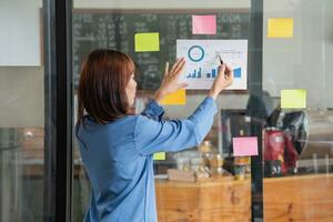 un' femmina dipendente scrive su un' colorato bloc notes. etnico donne Lavorando a startup brainstorming collaborativo piani su bicchiere parete adesivi vedere meno. foto