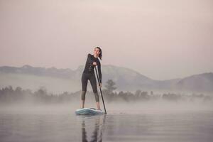 donna In piedi su paddling su lago Kirchsee a mattina nebbia, cattivo toelz, Baviera, Germania foto