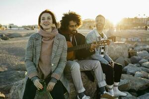 tre contento amici con chitarra seduta all'aperto a tramonto foto