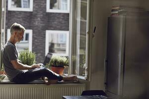maturo uomo seduta a il finestra a casa utilizzando il computer portatile foto